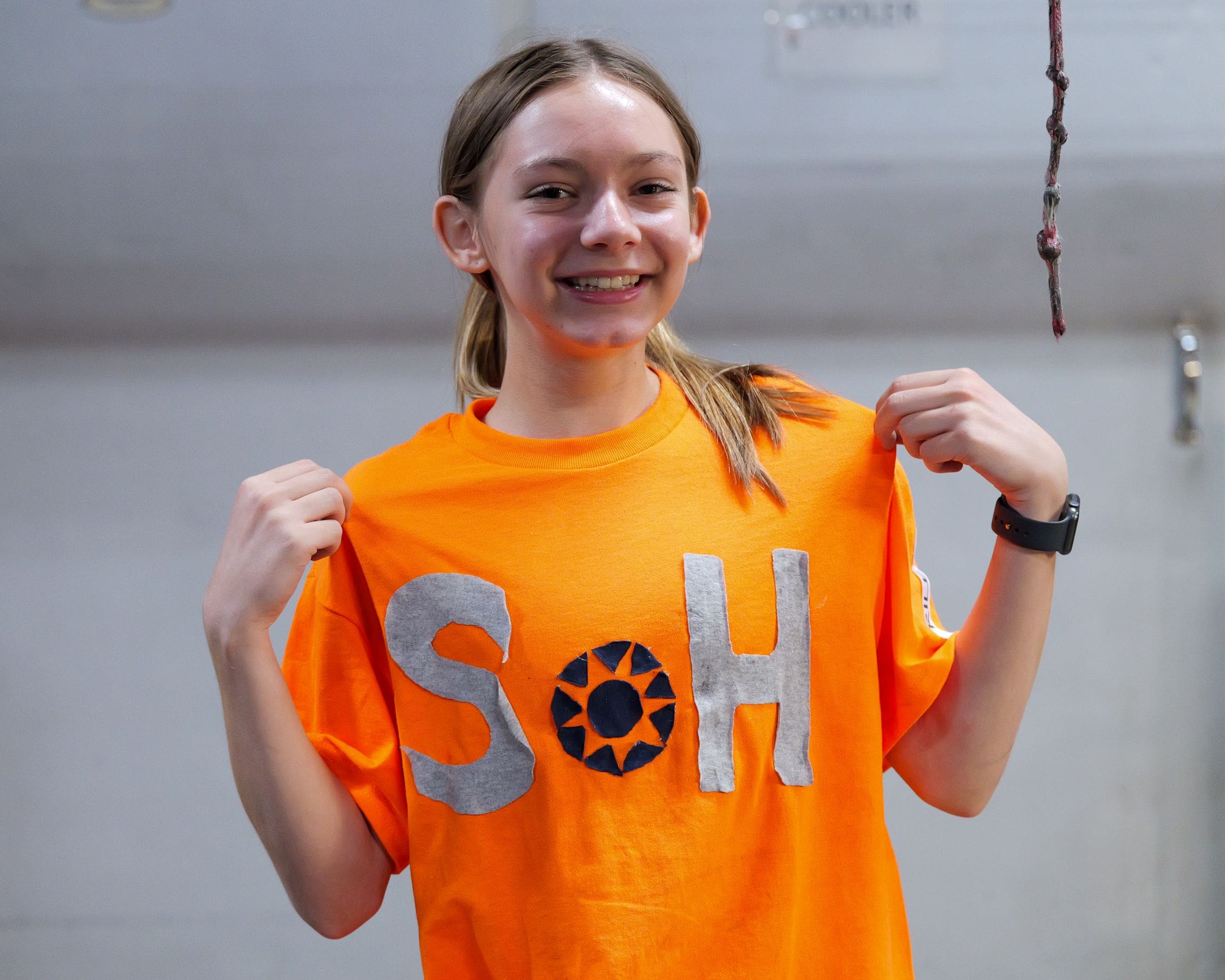 Smiling girl in an orange t shirt with fists raised a rope hanging in the background in a gym like setting