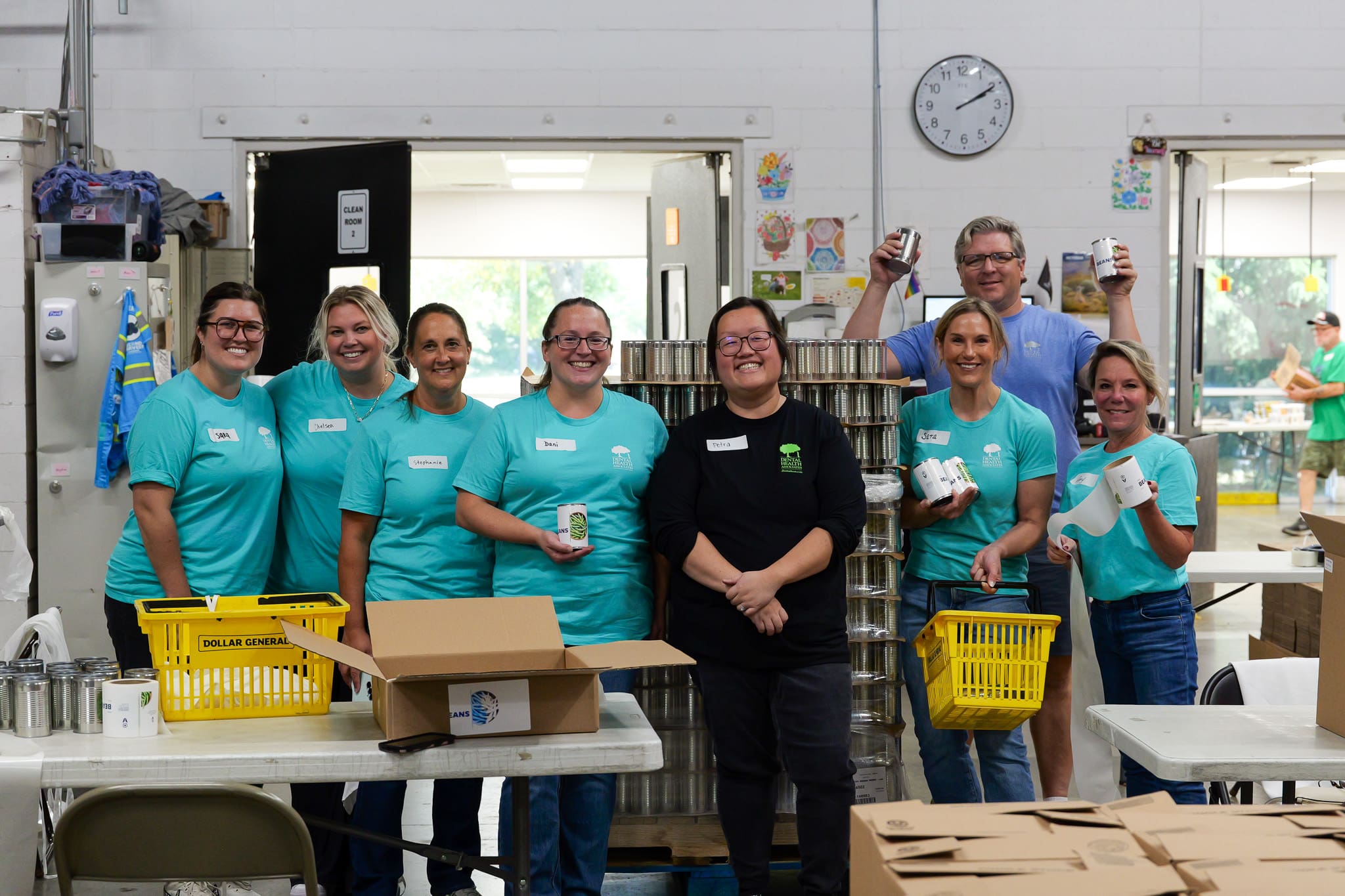 Group of smiling volunteers in teal shirts standing behind tables with canned goods in a warehouse setting holding up cans and boxes
